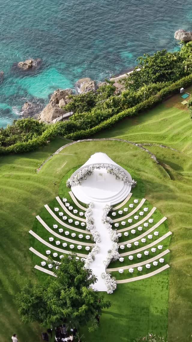 Aerial view of a white wedding ceremony setup with floral aisle and altar overlooking the ocean.