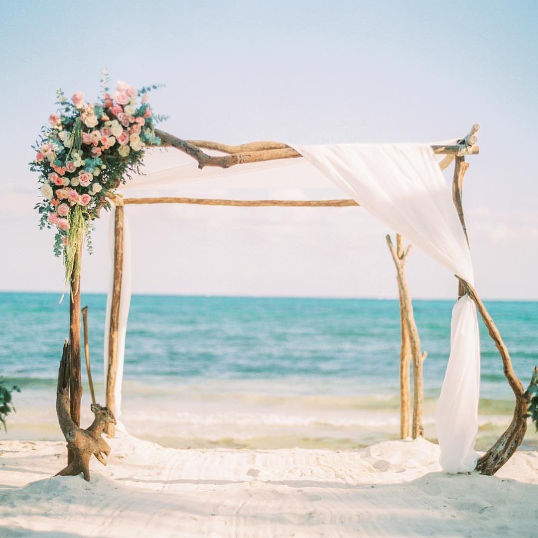Driftwood wedding altar with white fabric and pink flowers on a beach.