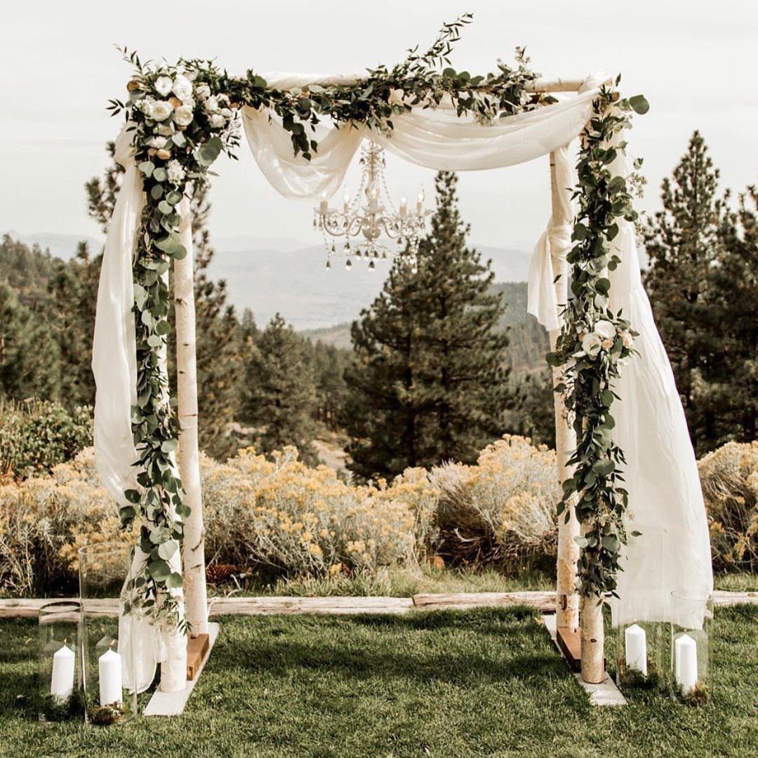 Birch pole archway decorated with white fabric, greenery, white roses, and a hanging crystal chandelier.