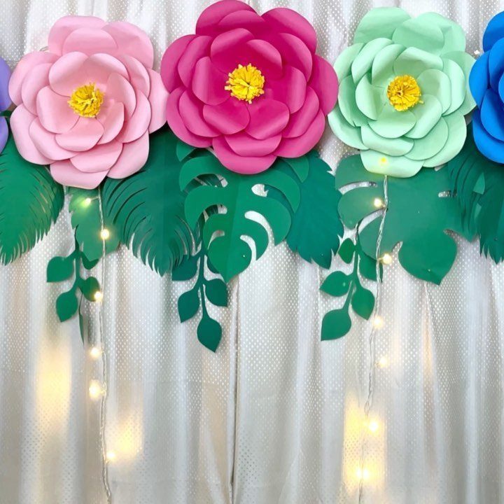 Large colorful paper flowers and green leaves decorating a white fabric backdrop with fairy lights.