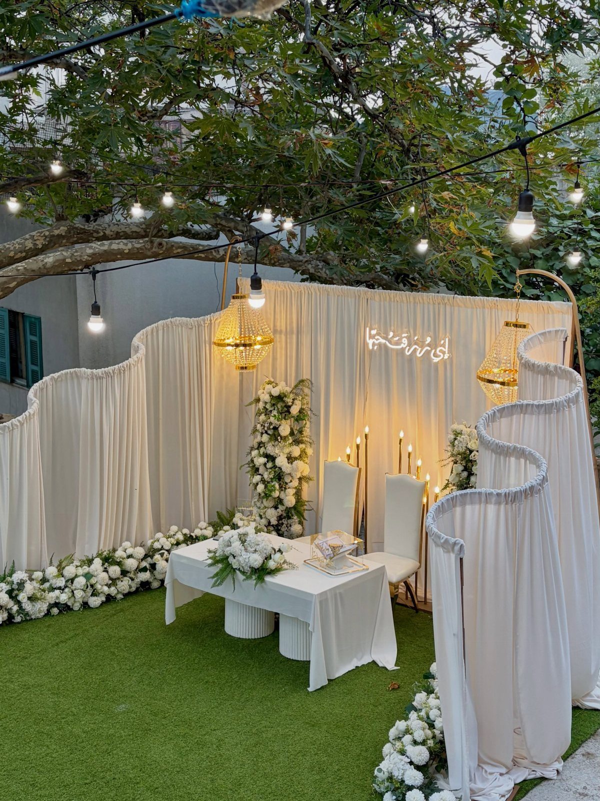 White draped fabric backdrops with floral arrangements and gold chandeliers on green grass.