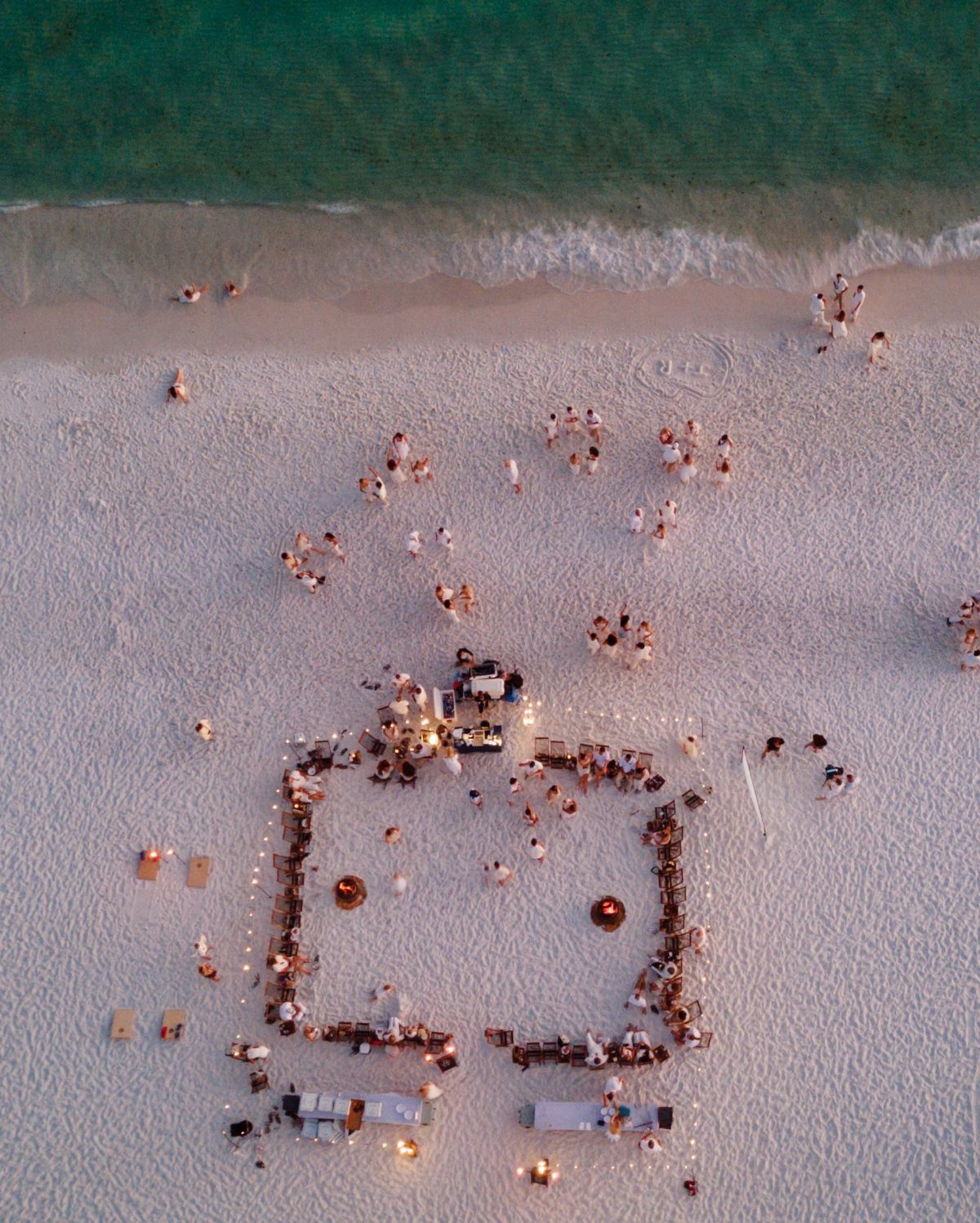 Aerial view of people gathered around a square arrangement of fires on a sandy beach.
