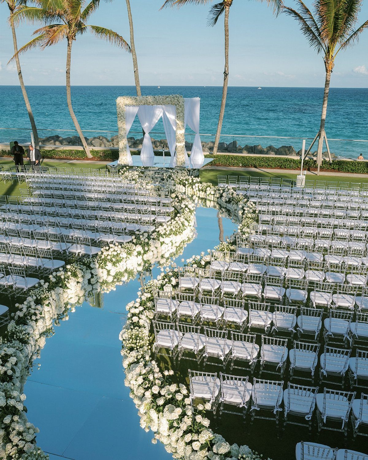 White wedding chairs arranged around a winding blue aisle runner decorated with white flowers by the ocean.