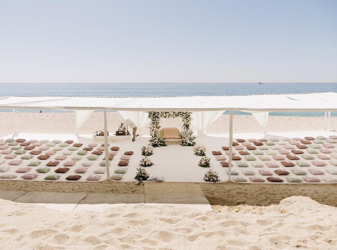 Rows of pastel cushions arranged on a sandy beach under a white canopy for wedding guests.