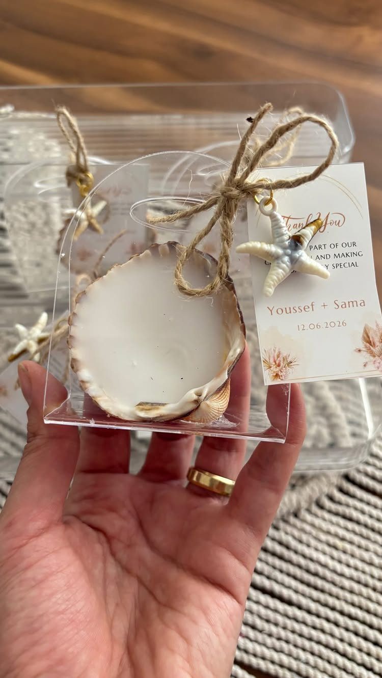 A hand holding a clear box containing a seashell candle and a starfish charm.