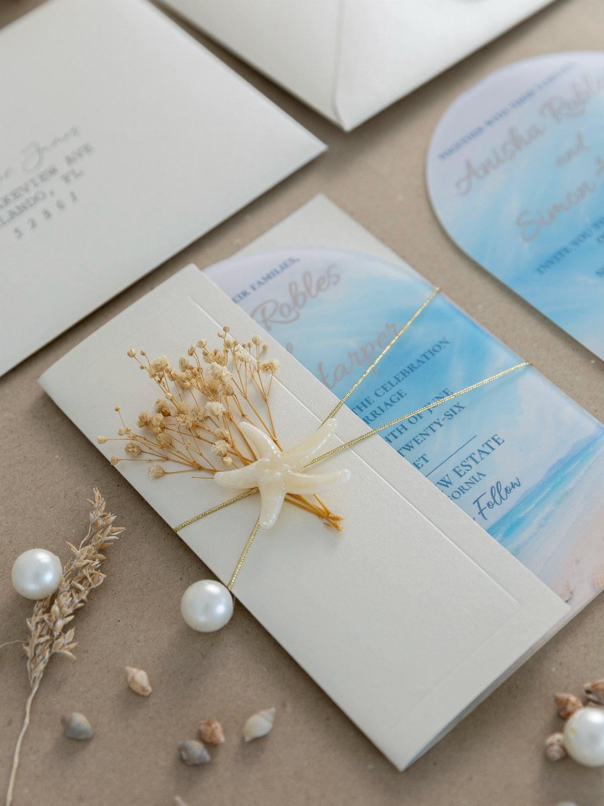 Beach-themed wedding invitations with blue watercolor patterns, starfish, pearls, and dried flowers.