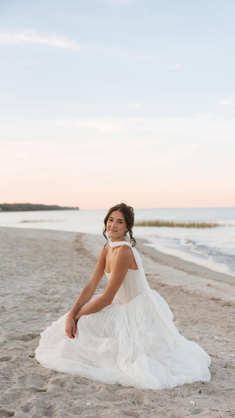 Woman in a flowing white wedding dress sitting on a sandy beach at sunset.