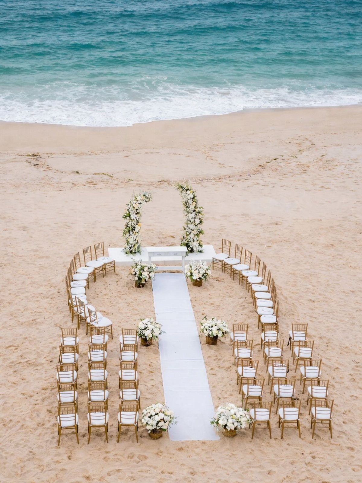 White wedding ceremony setup with floral pillars and gold chairs on a sandy beach.