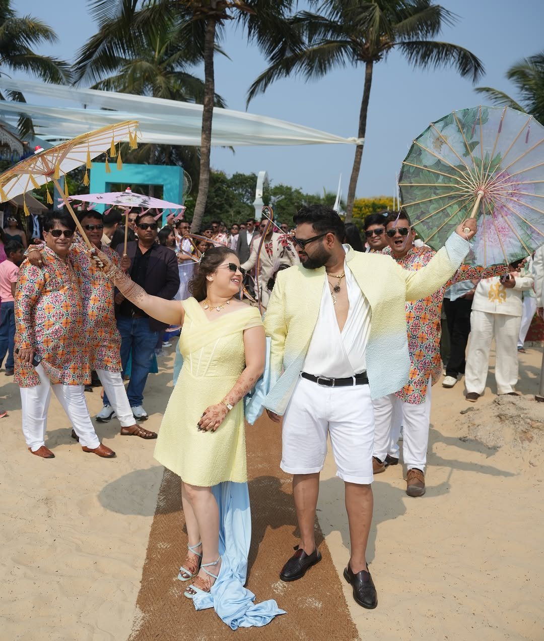 A man in a yellow blazer and a woman in a yellow dress walking on a beach.
