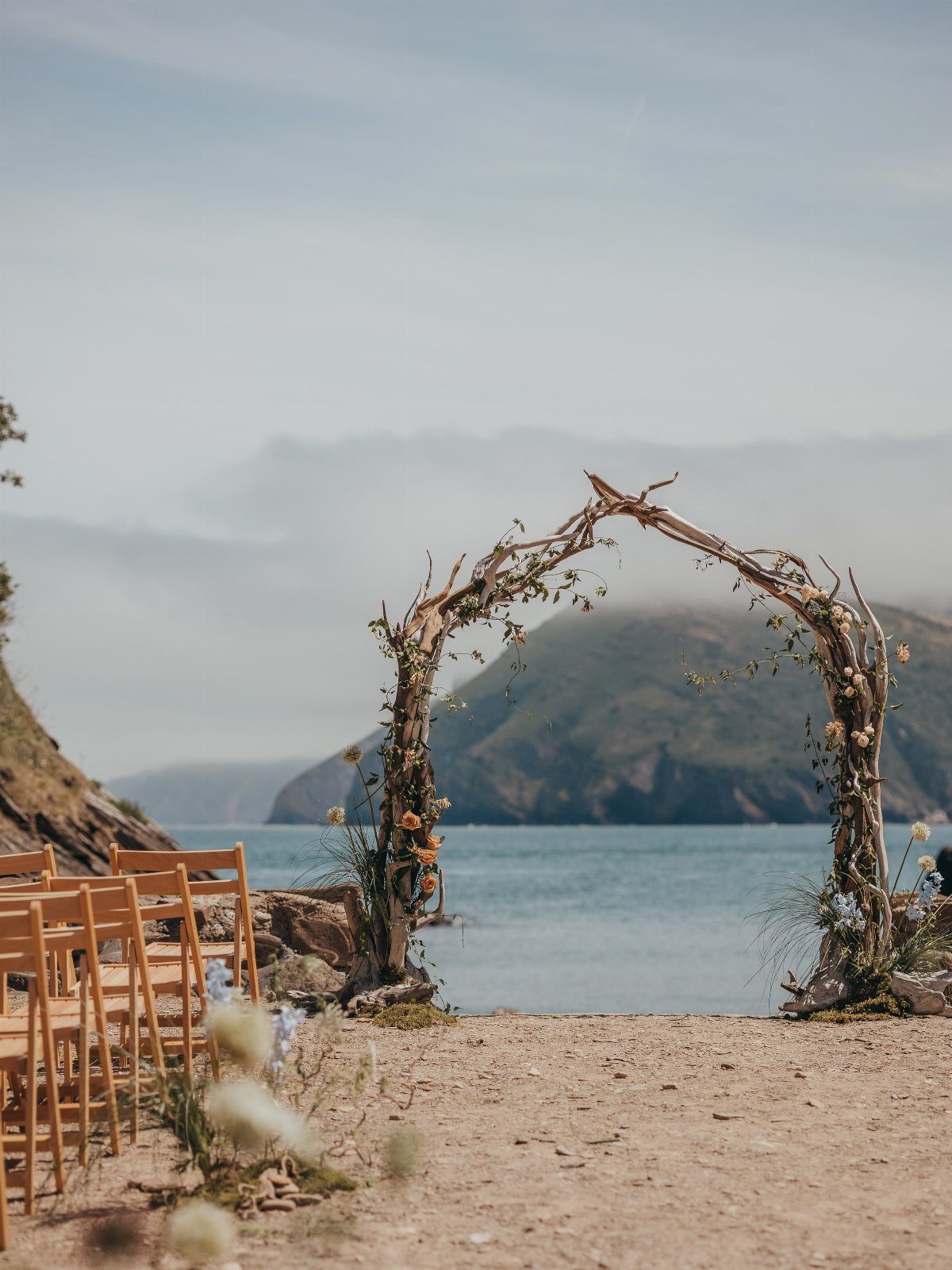 A driftwood ceremony arch decorated with flowers stands on a beach overlooking the ocean.