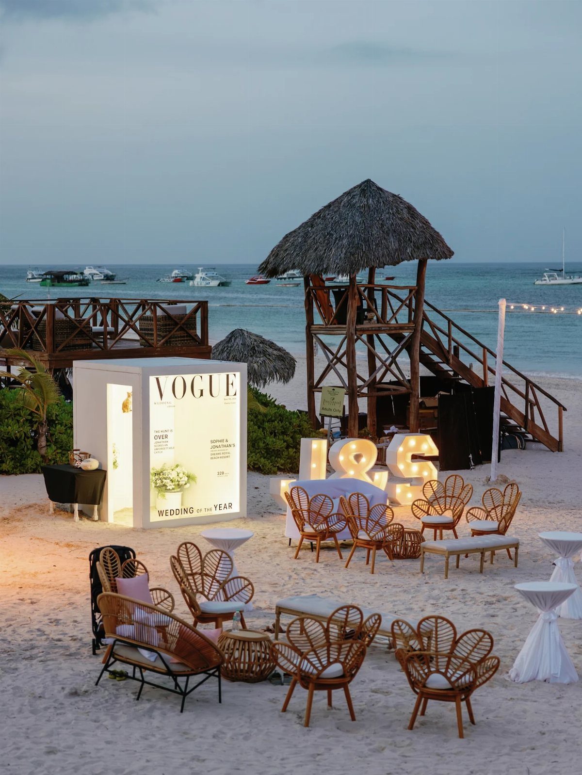 Wicker chairs and illuminated marquee letters arranged on a sandy beach near the ocean at dusk.