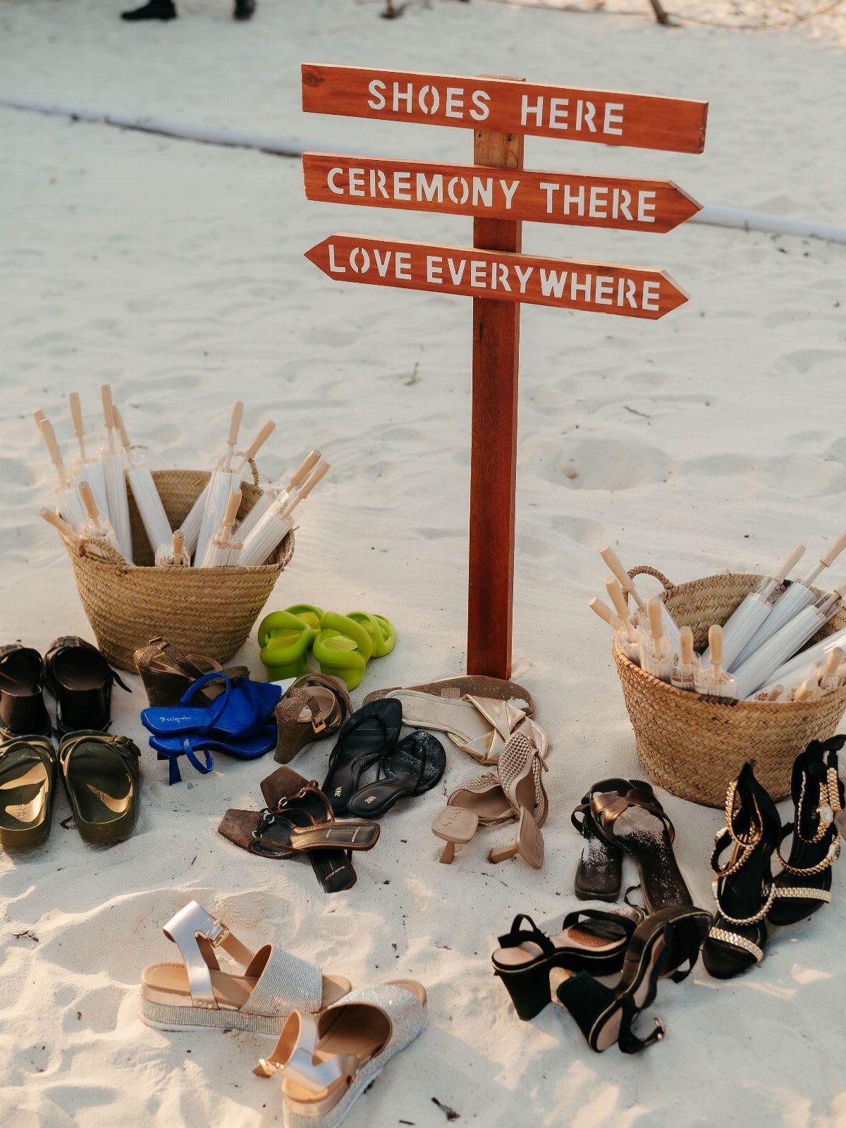 Wooden sign on sand surrounded by various women's sandals and baskets of flip-flops.