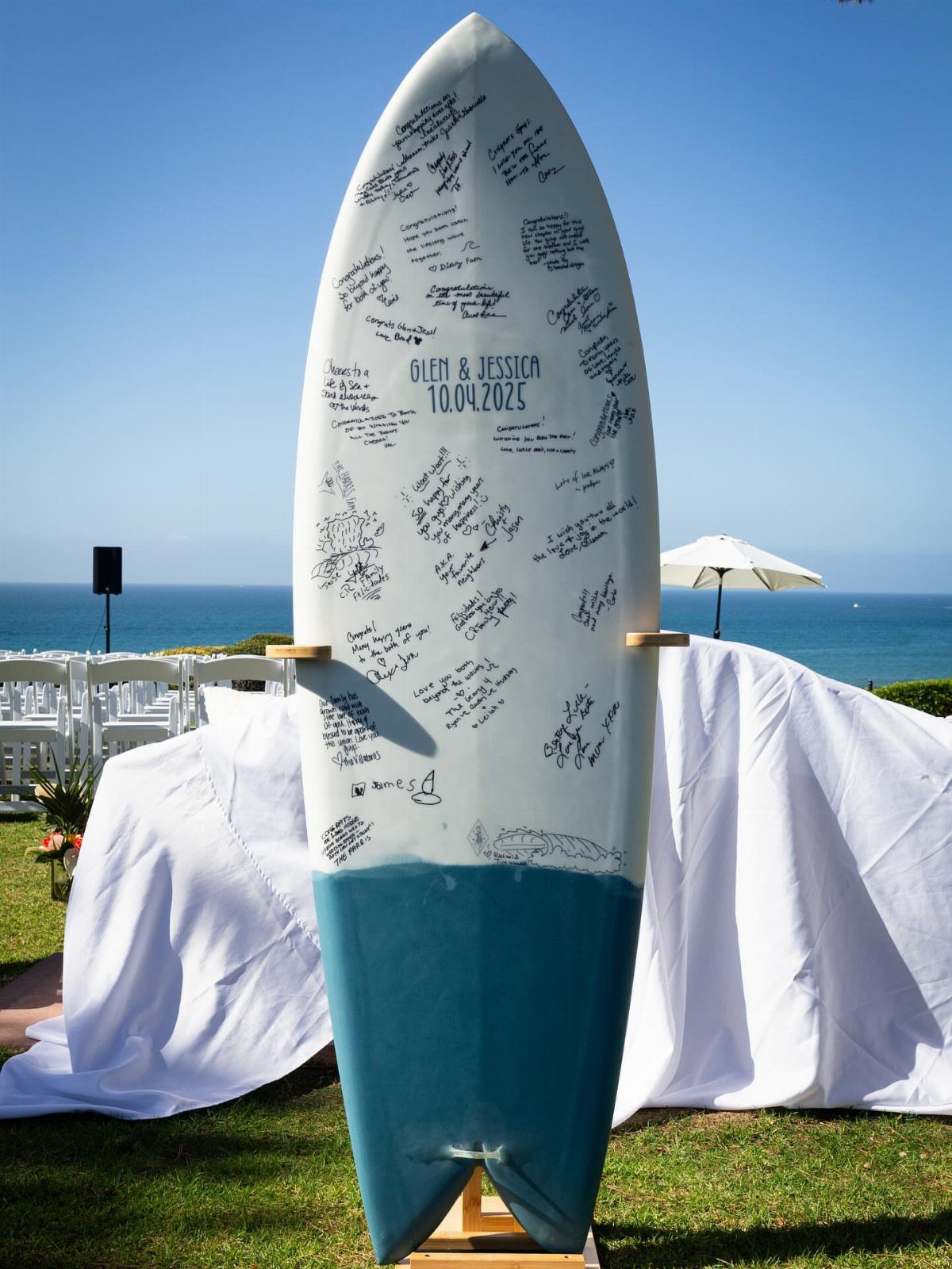 A white and teal surfboard covered in handwritten guest messages stands on an easel by the ocean.