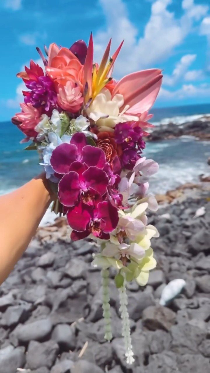 A hand holding a vibrant tropical bouquet of orchids and lilies by the ocean.