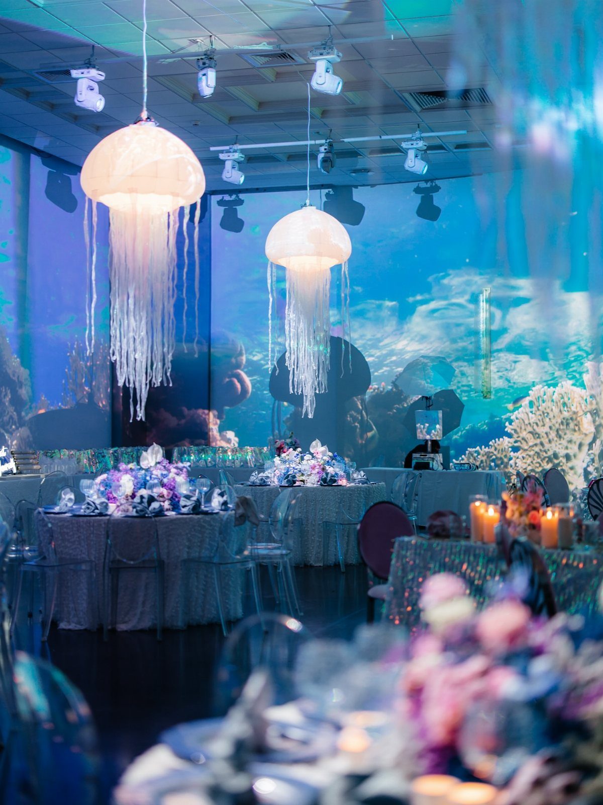 Glowing jellyfish hanging lamps over decorated tables in an underwater themed event space.