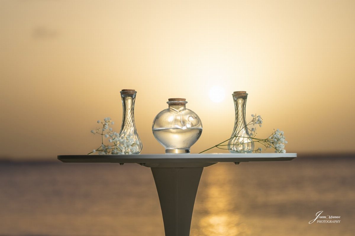 Three glass vessels with water and white flowers on a table at sunset.
