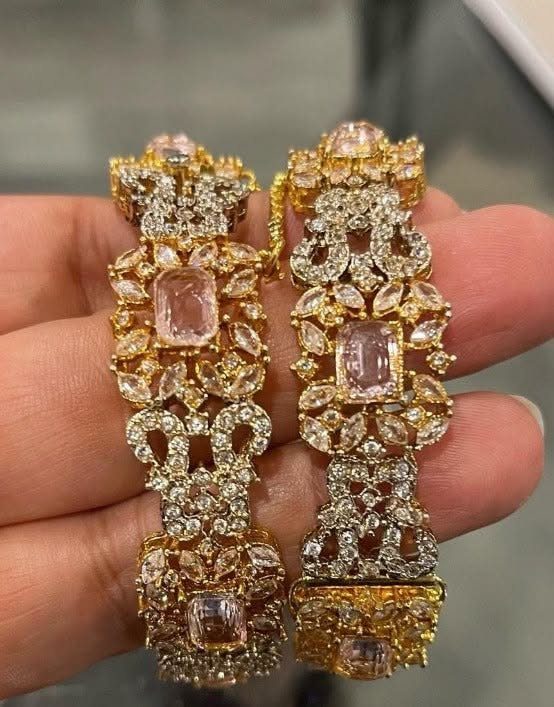 Two wide gold bangles adorned with pink and clear gemstones held in a hand.