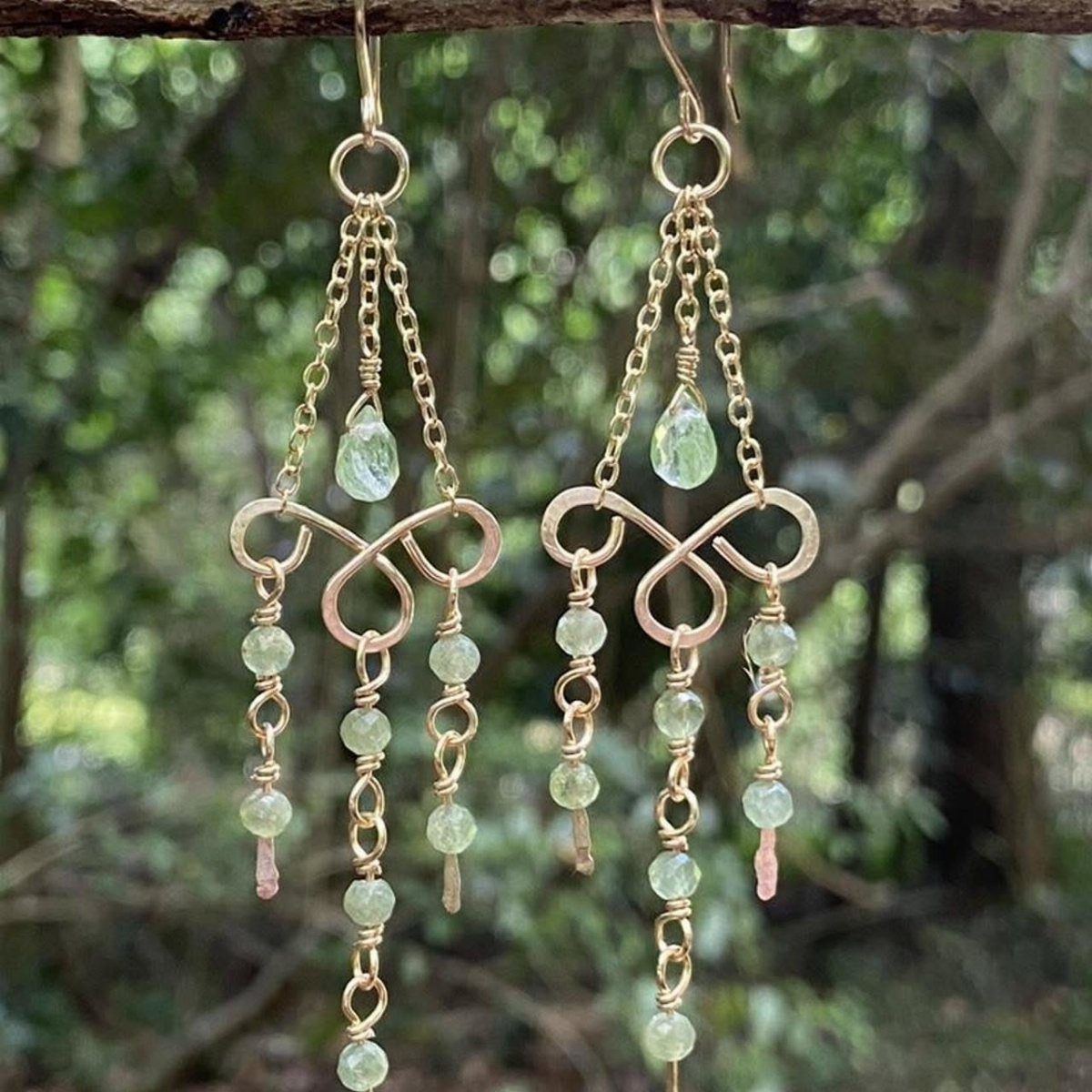 Gold chandelier earrings with light green gemstones hanging against a blurred green forest background.
