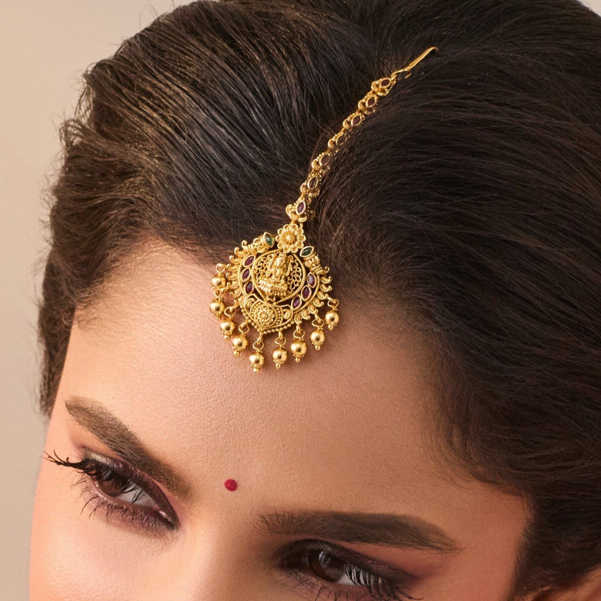 Gold maang tikka headpiece with intricate carvings and dangling beads worn on a woman's forehead.