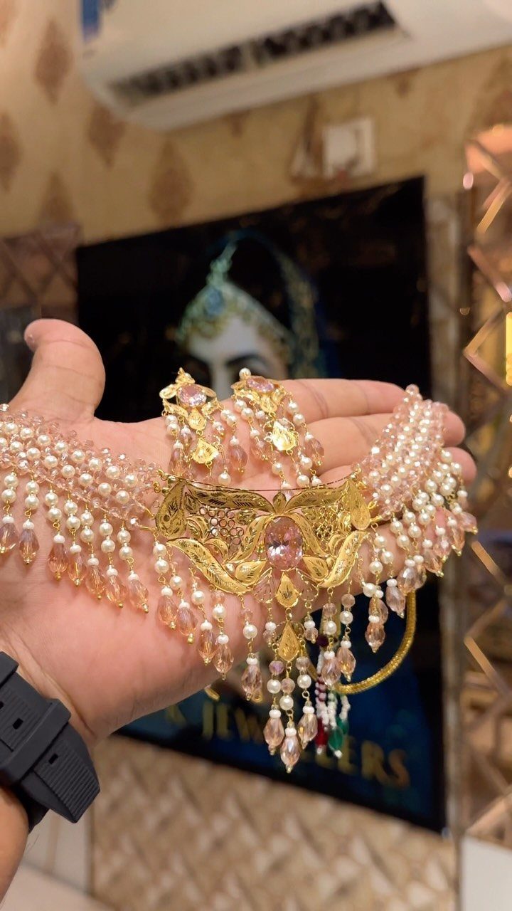 A hand holding an intricate gold necklace adorned with many small pearls and peach gemstones.