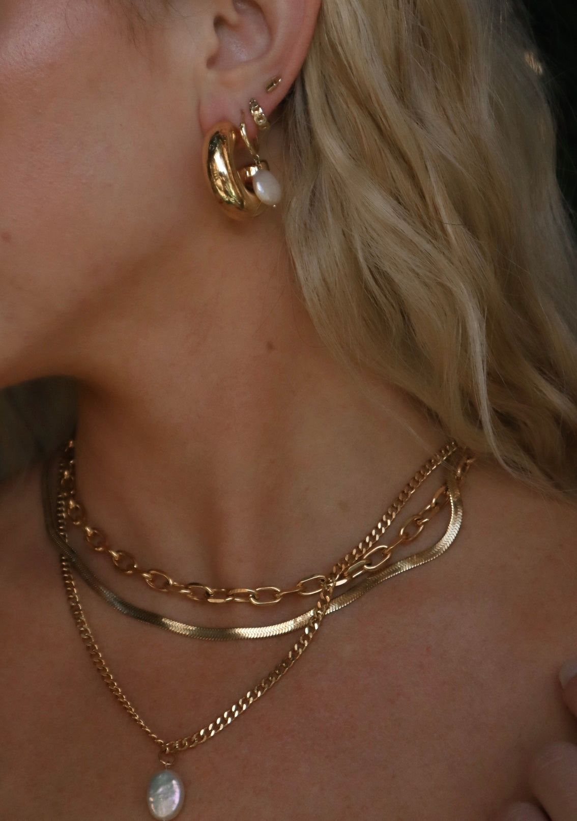 A woman wearing layered gold necklaces with a pearl pendant and gold hoop earrings.