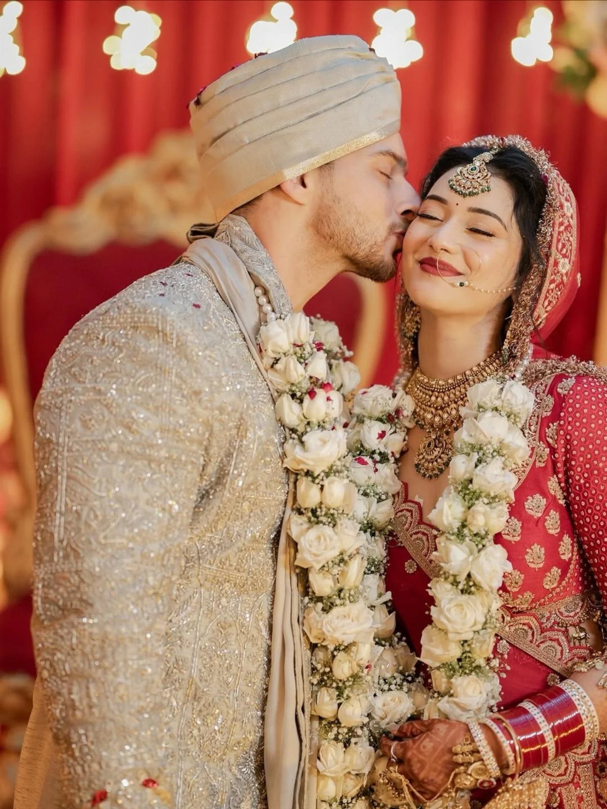 A groom in traditional attire kisses a smiling bride wearing red wedding clothing and garlands.