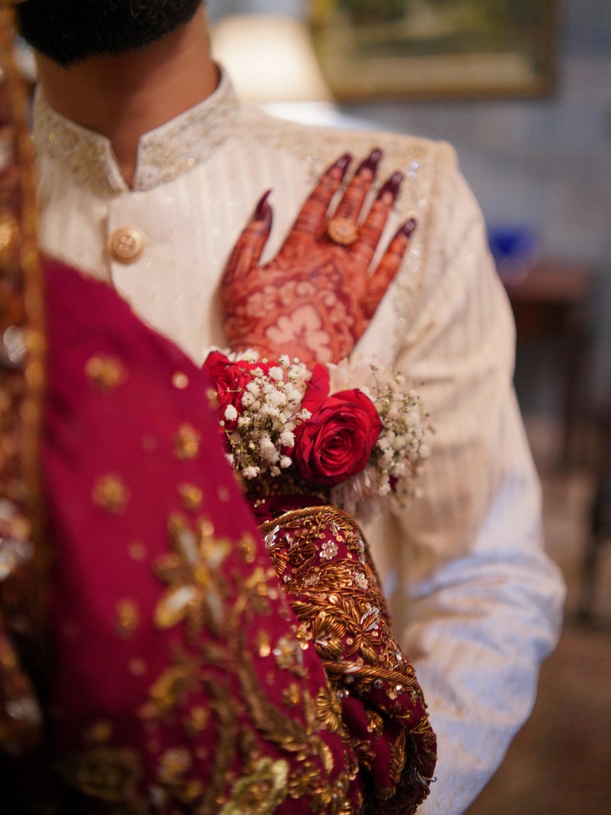 A hand with henna patterns rests on a cream sherwani next to red roses.
