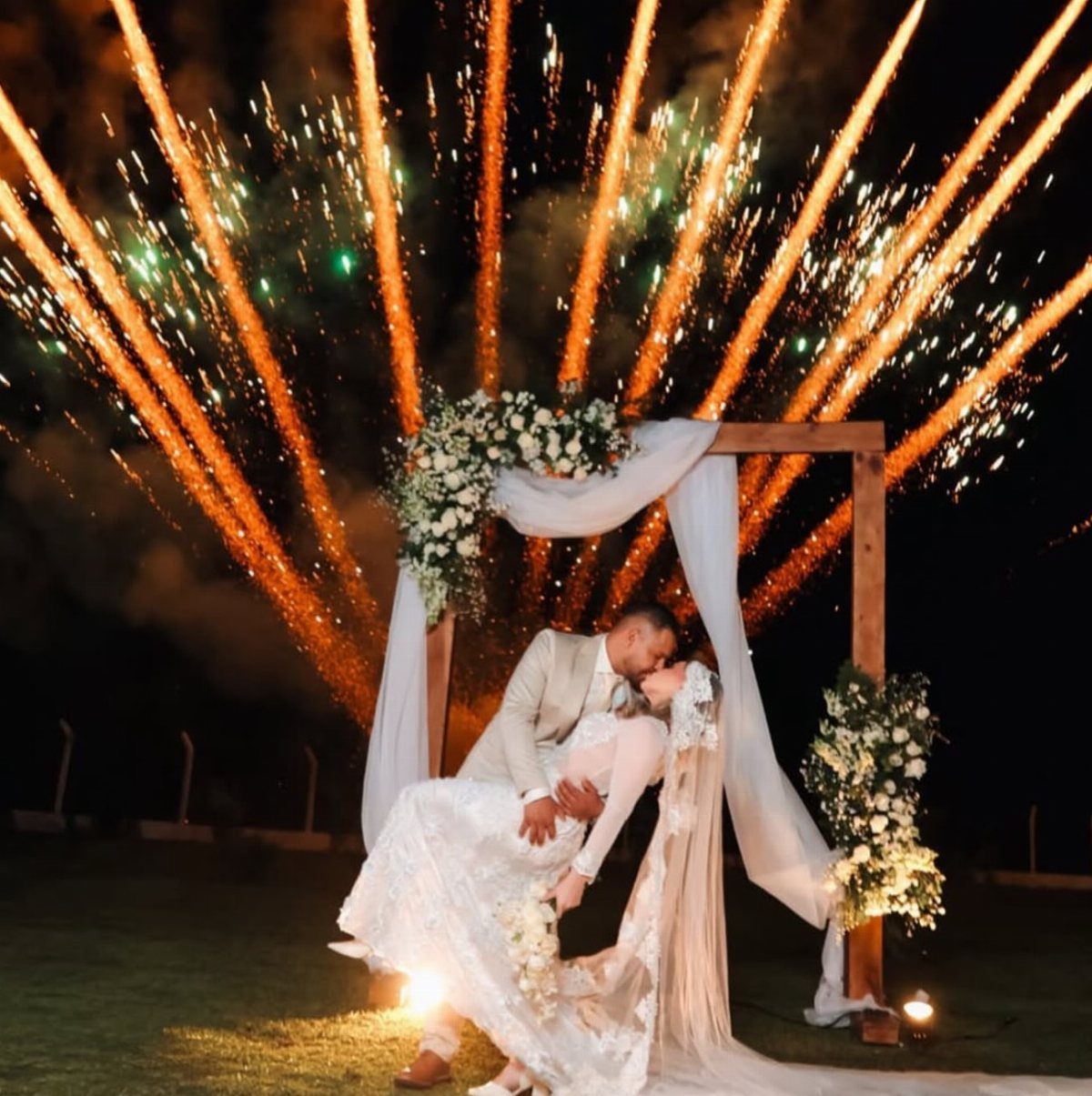 A bride and groom embracing under a floral wedding arch with fireworks exploding in the background.