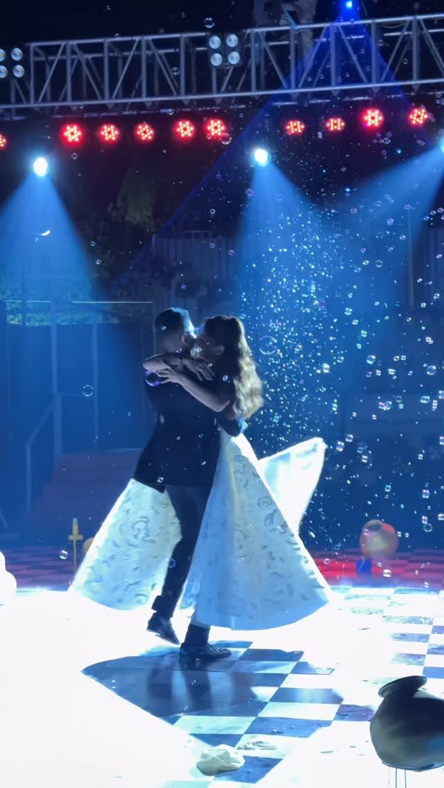 A bride and groom dancing together on a checkered floor under blue stage lights and bubbles.