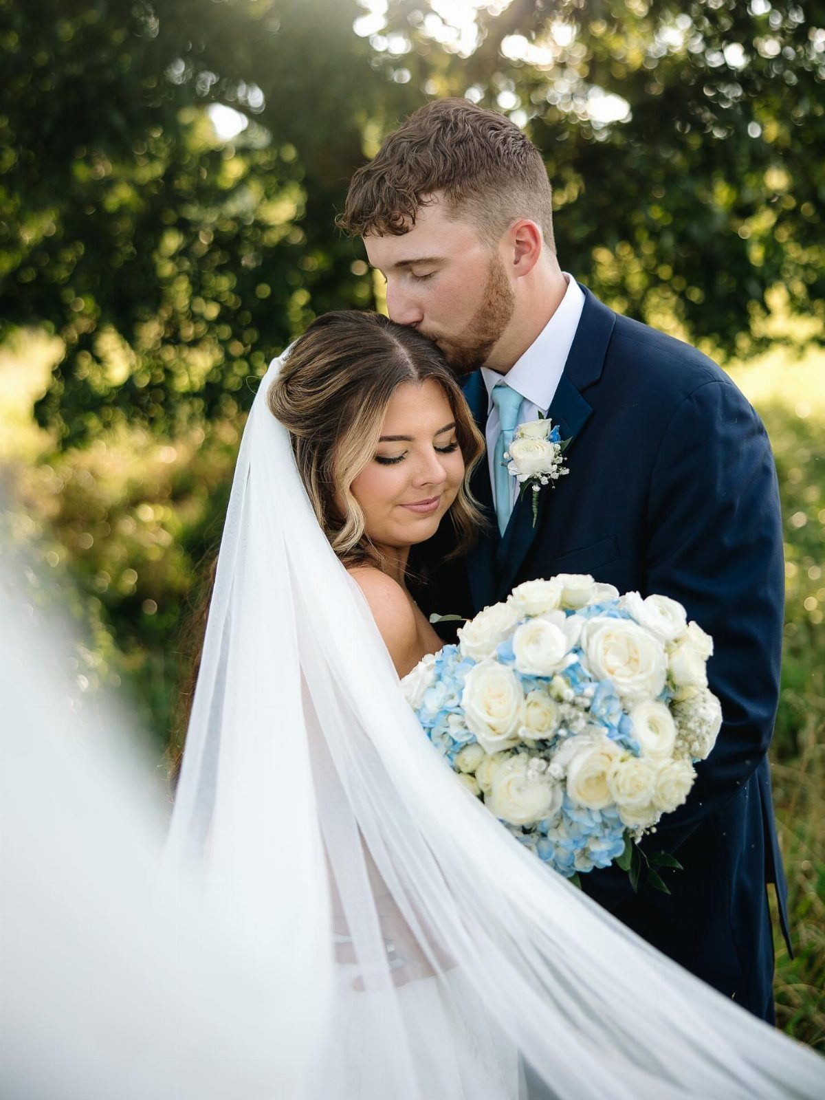 A groom kisses the bride's forehead while she holds a white and blue rose bouquet.