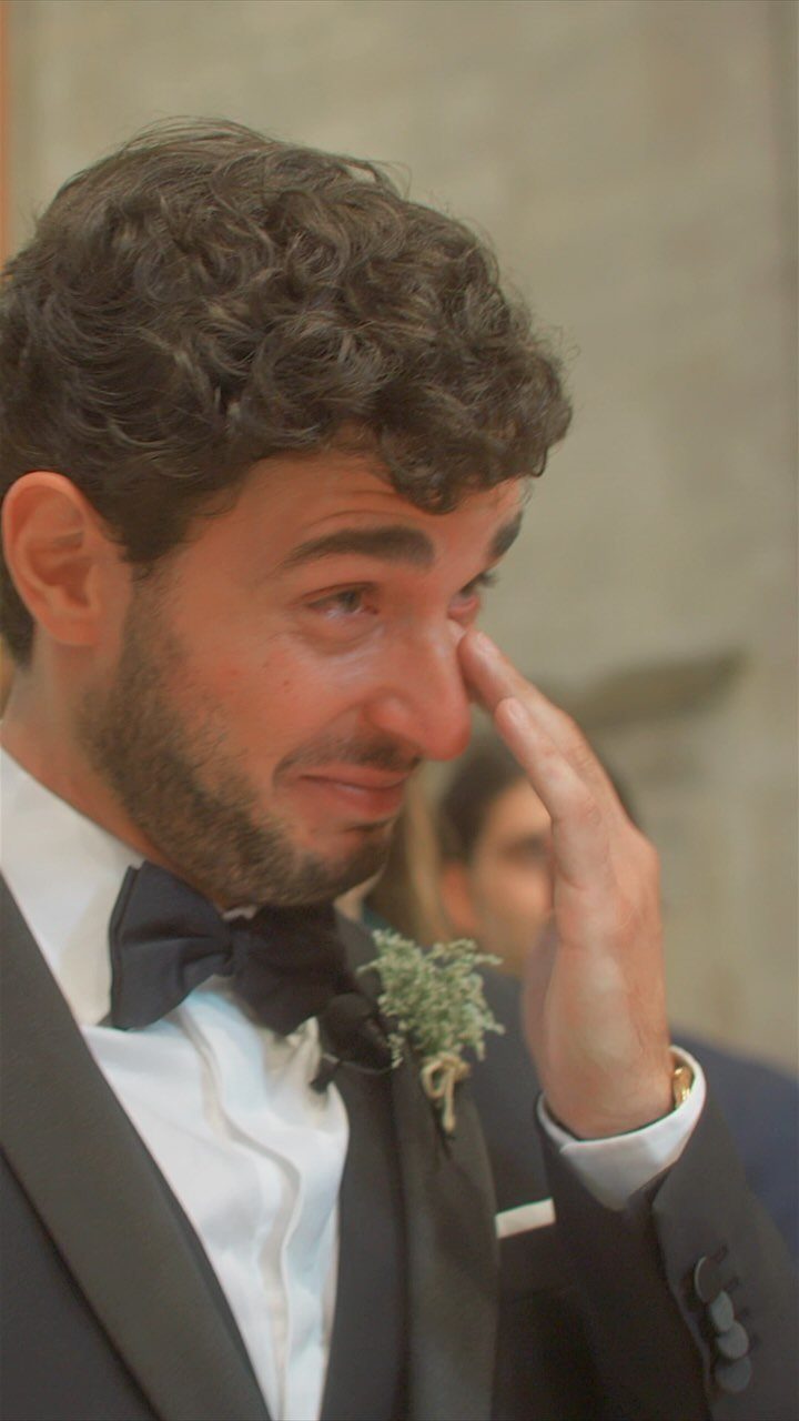 A groom in a black tuxedo wipes away a tear during a wedding ceremony.