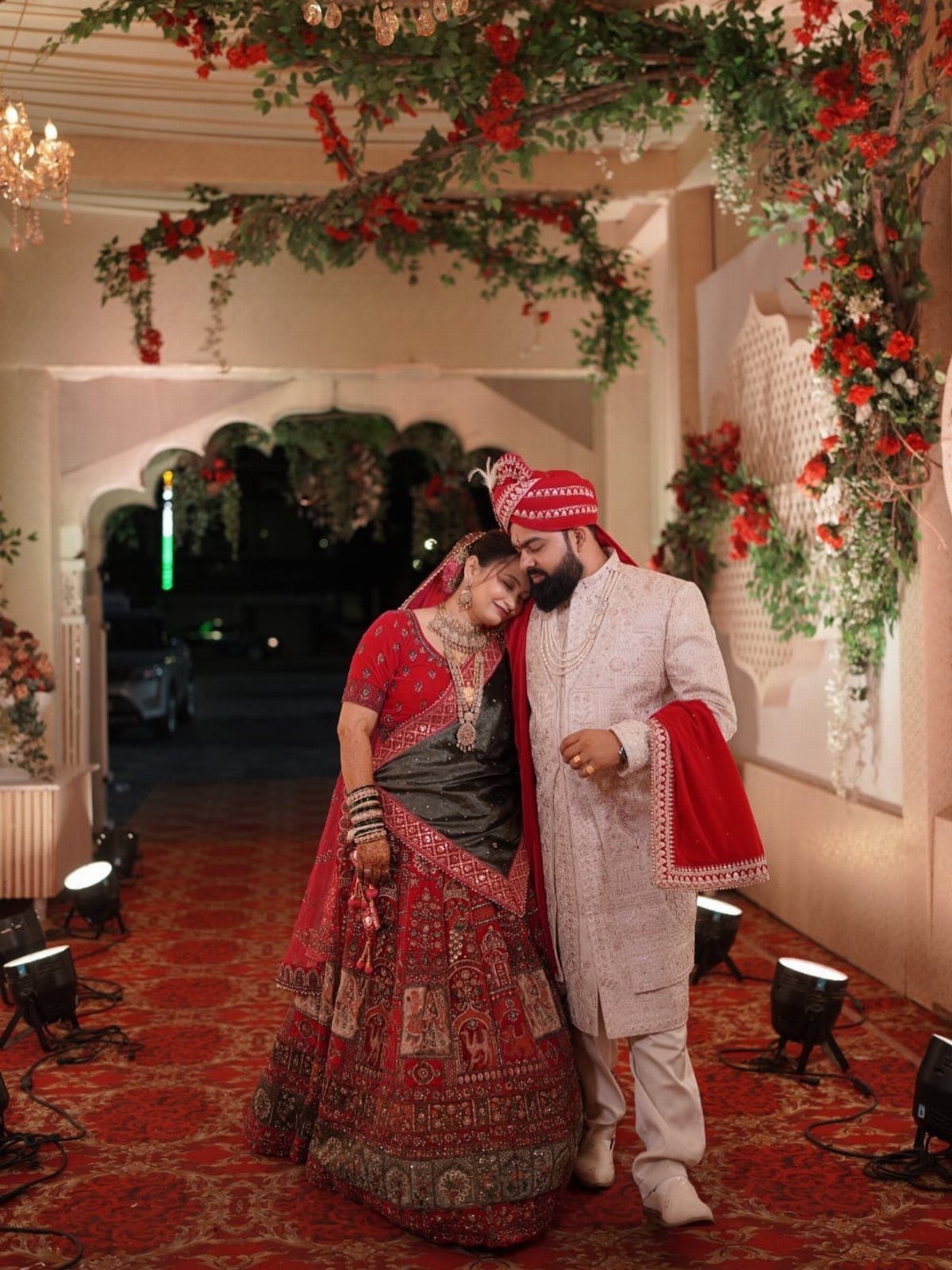 Bride in red lehenga leaning on groom in white sherwani under floral arch.
