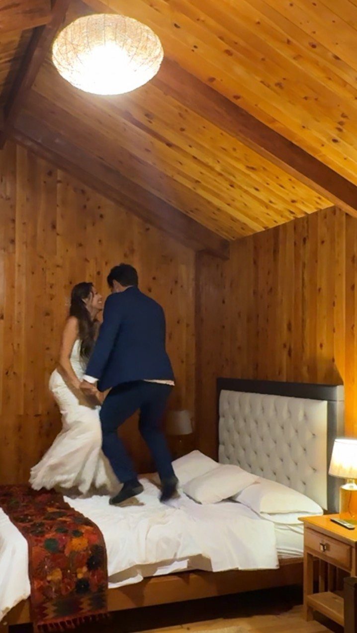 Bride and groom in wedding attire jumping onto a bed in a wooden room.
