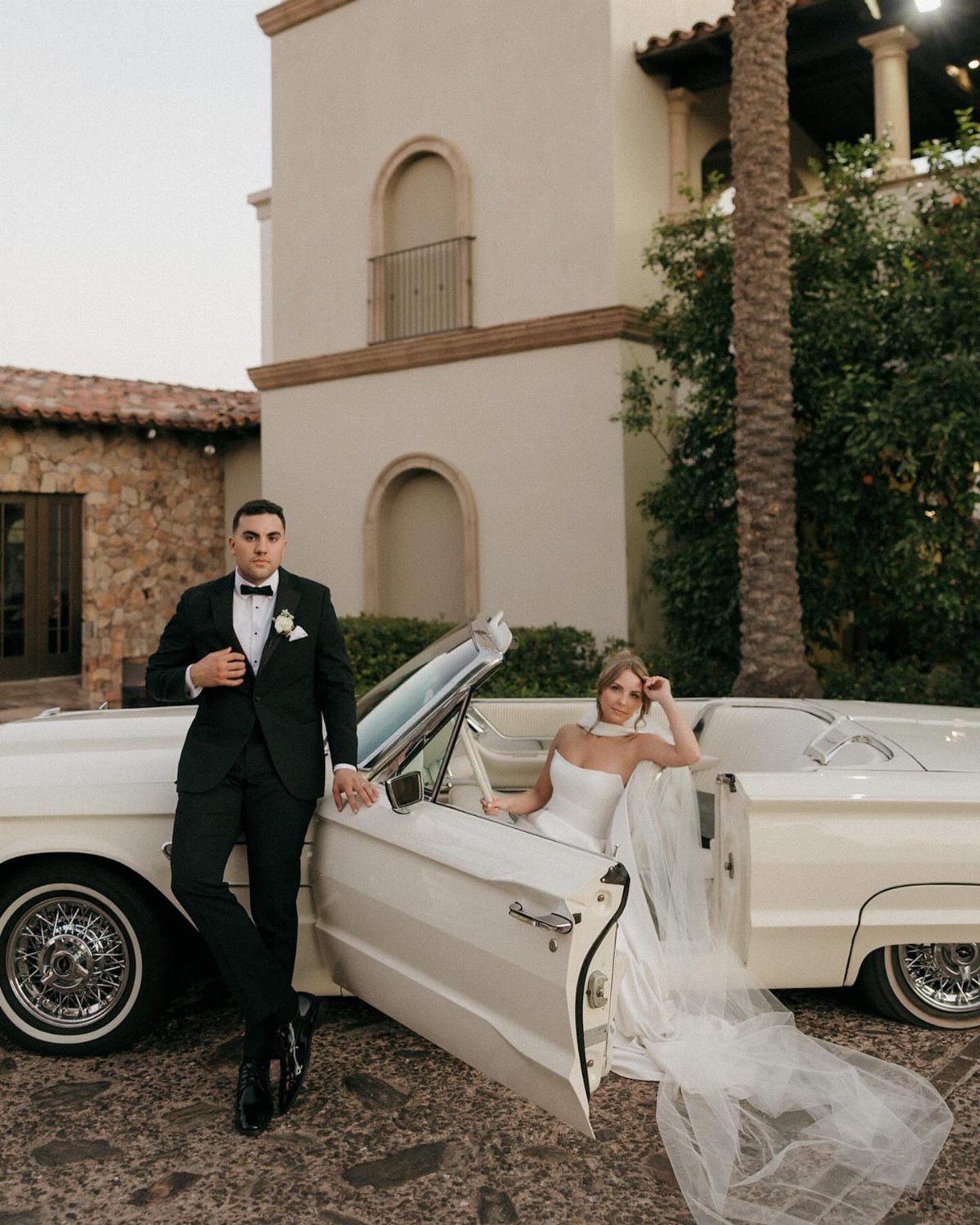 A bride in a white dress sits in a vintage car while the groom stands beside it.