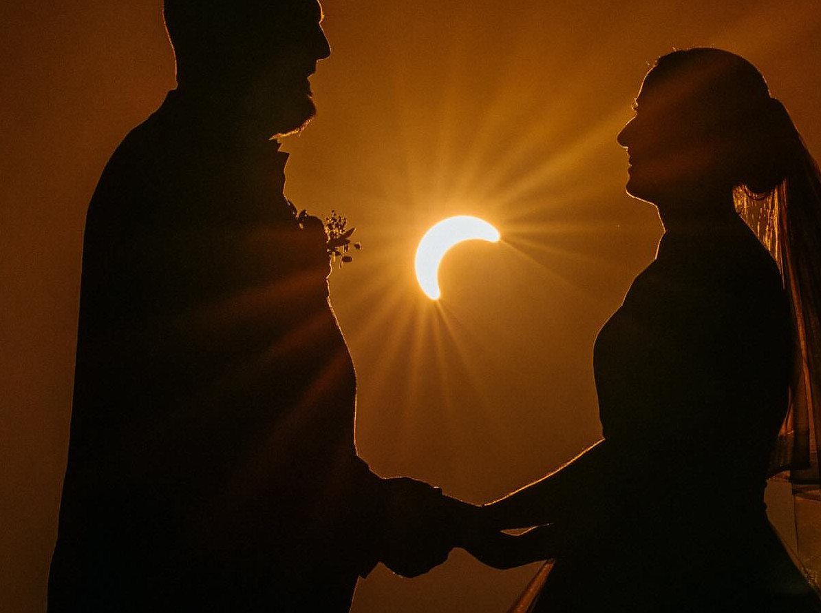 Silhouetted bride and groom holding hands against a bright orange sunset with a crescent sun.