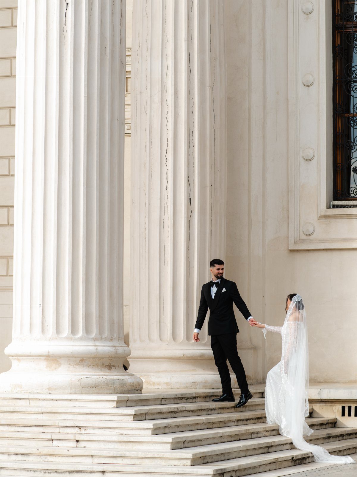 Bride and groom walking down white marble steps beside large classical columns.