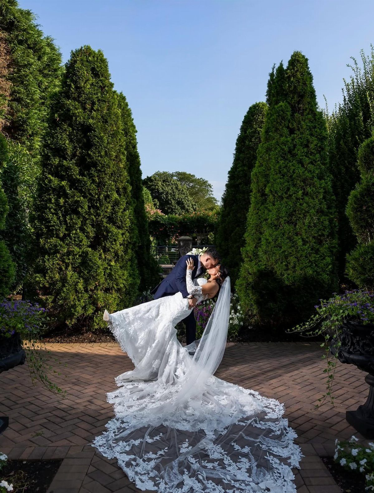 A bride and groom embracing on a brick path surrounded by tall green evergreen trees.