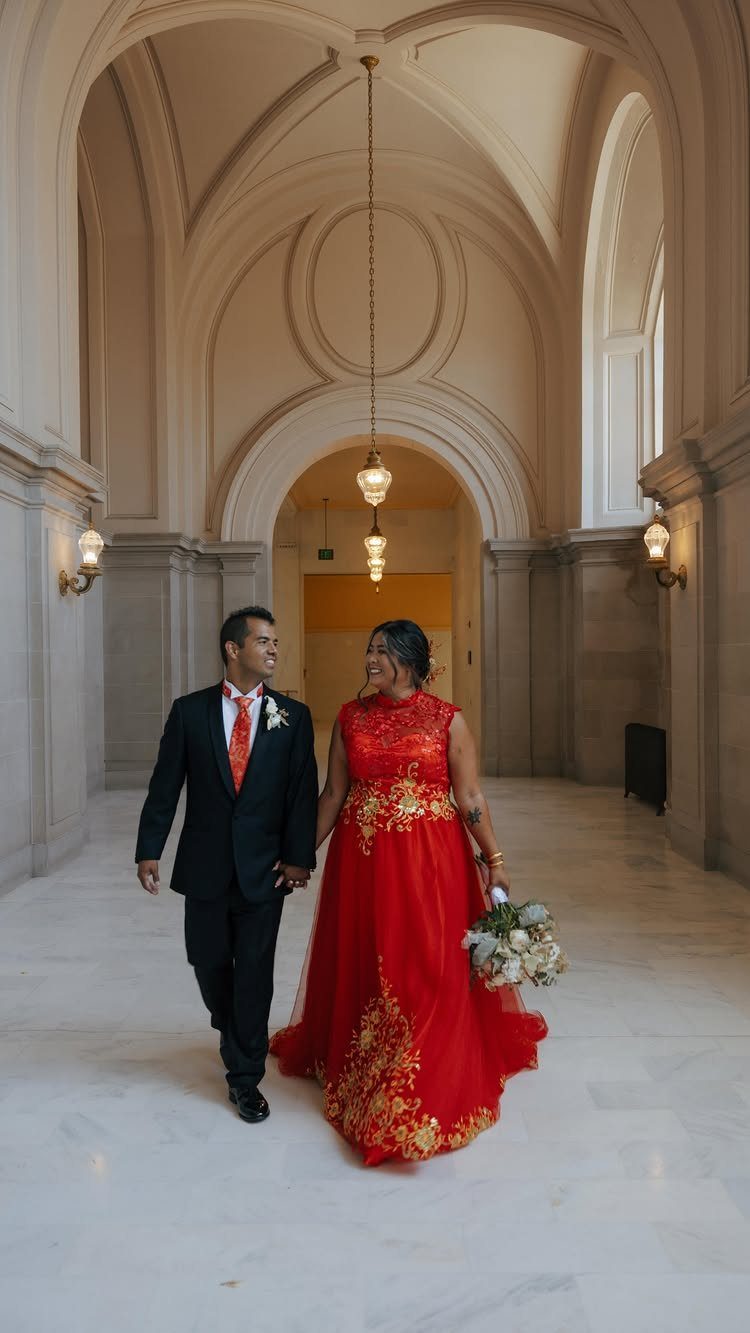 A bride in a red embroidered gown and a groom in a black suit walk through an arched hallway.