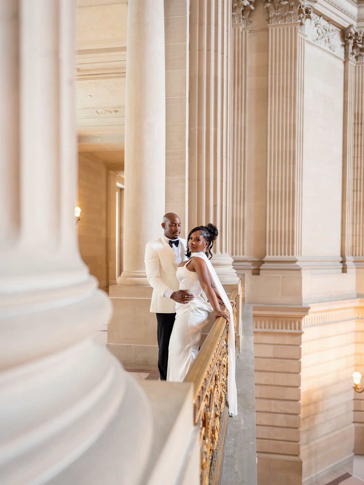 A bride in a structured white gown and a groom in a white tuxedo pose on a grand staircase.