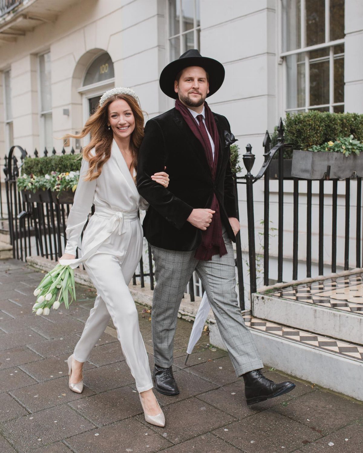 Woman in a white satin jumpsuit and pearl headband walking with a man on a city street.