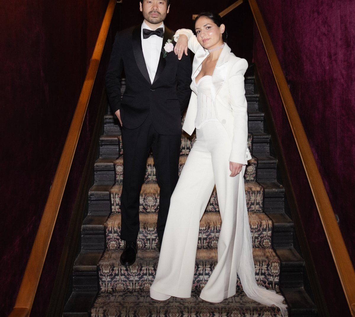 A couple posing on stairs, the woman wearing a white bridal pantsuit with a long train.