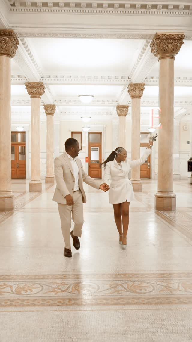 A man in a beige suit and a woman in a white blazer dress walking through a grand hall.