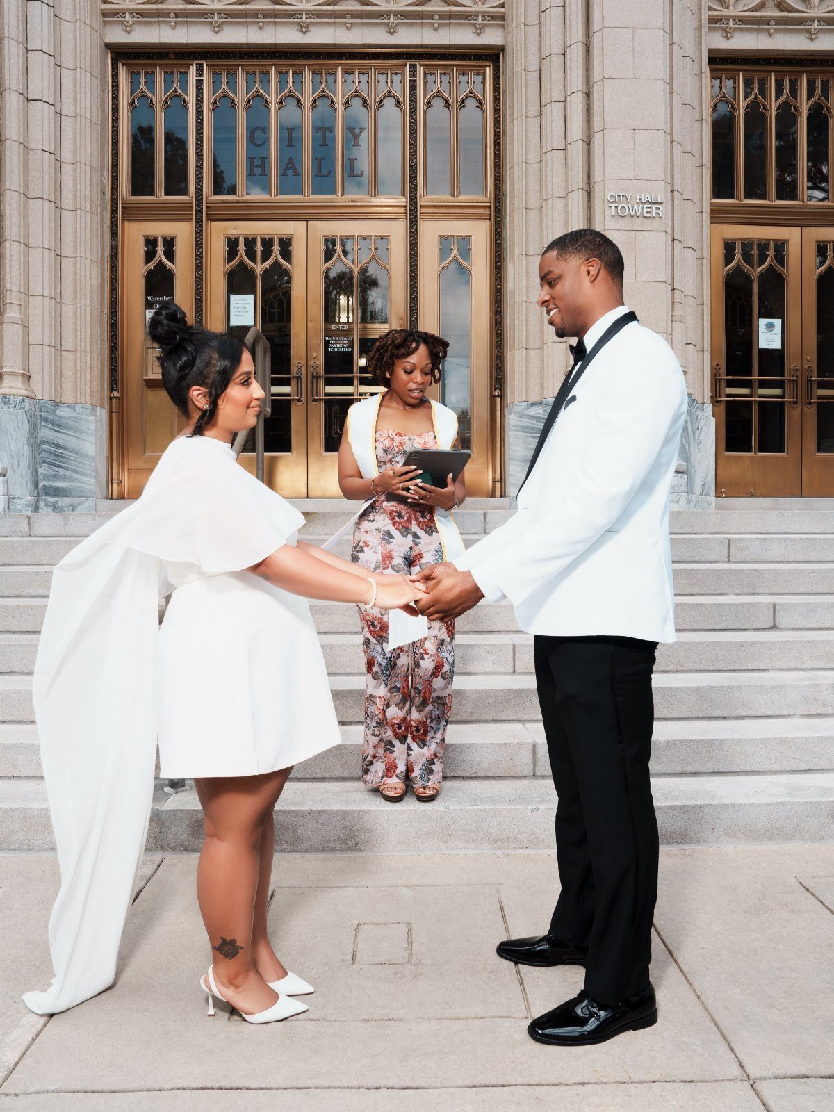 A bride in a short white cape dress and a groom in a white tuxedo jacket holding hands.