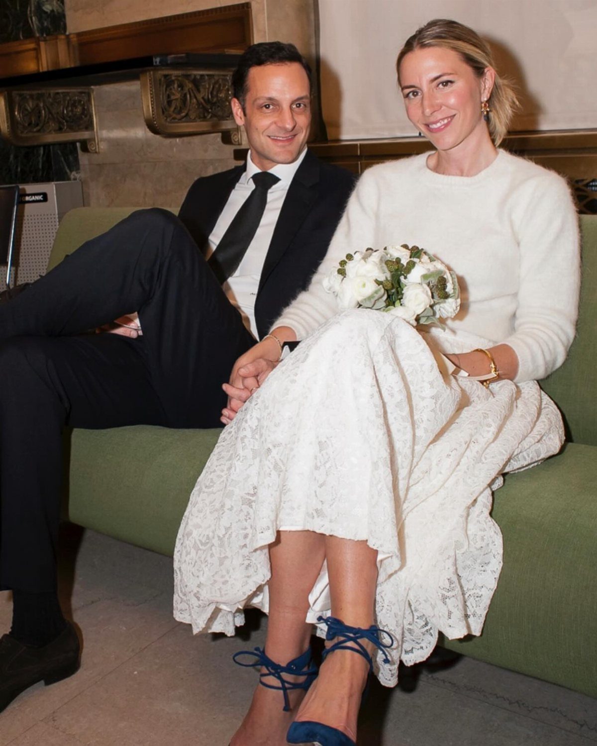 A bride in a white sweater and lace skirt holding flowers sits with a groom on a green sofa.