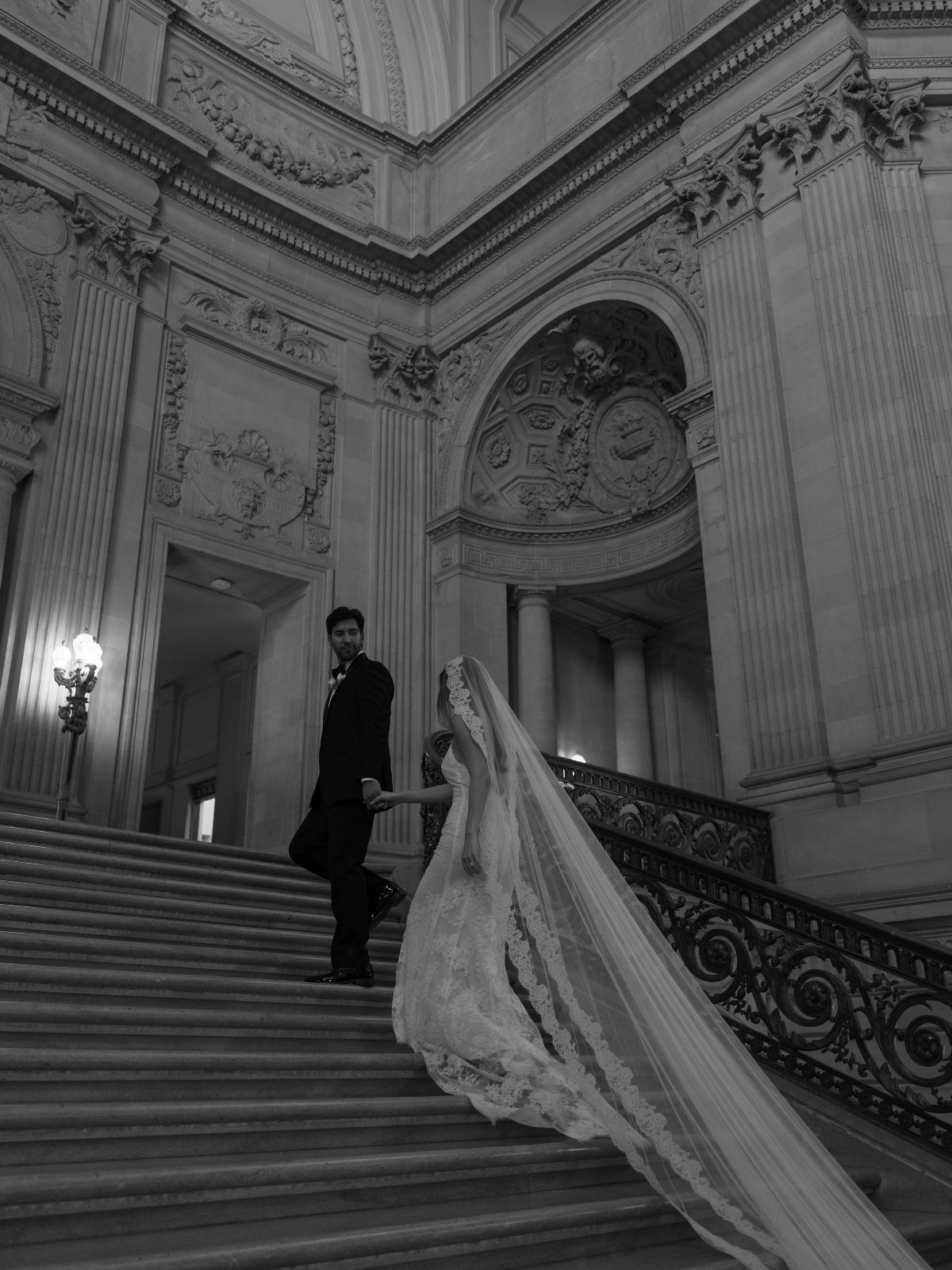 Bride in a lace wedding dress and long veil walking up grand stone stairs with her groom.