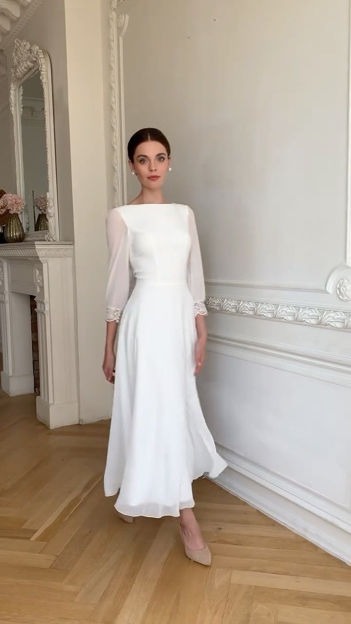 8 Civil Wedding Outfit Ideas for Brides Who Want Something Different 1 A woman posing in a long white wedding dress with sheer sleeves in a bright room.