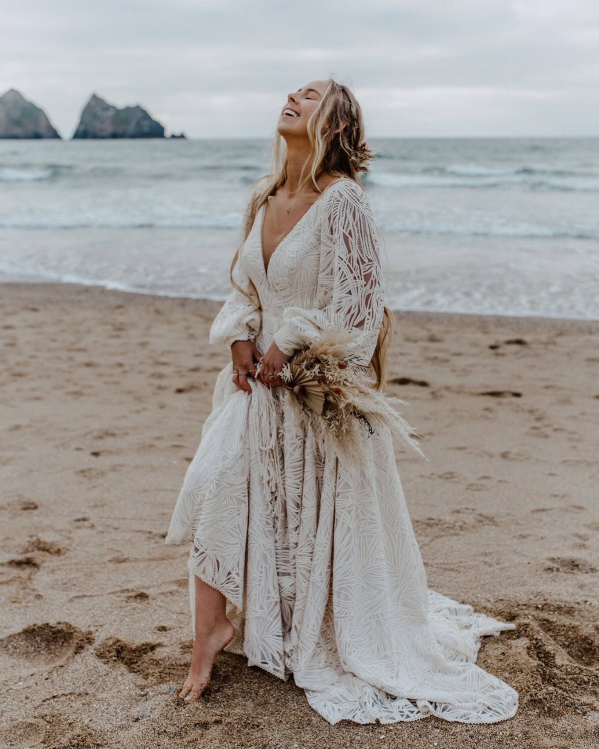 8 Civil Wedding Outfit Ideas for Brides Who Want Something Different 2 A woman in a white lace flowing dress stands on a beach holding a bouquet.