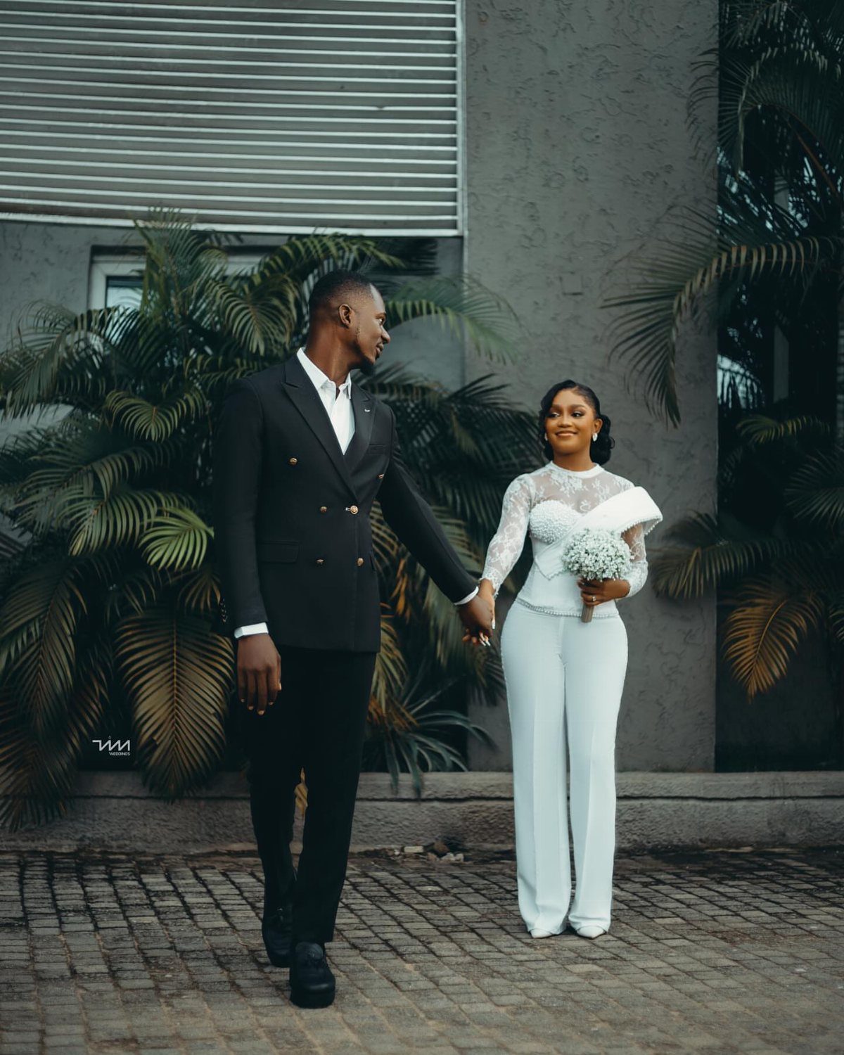 8 Civil Wedding Outfit Ideas for Brides Who Want Something Different 5 A man in a black suit and a woman in a white lace jumpsuit holding hands outdoors.