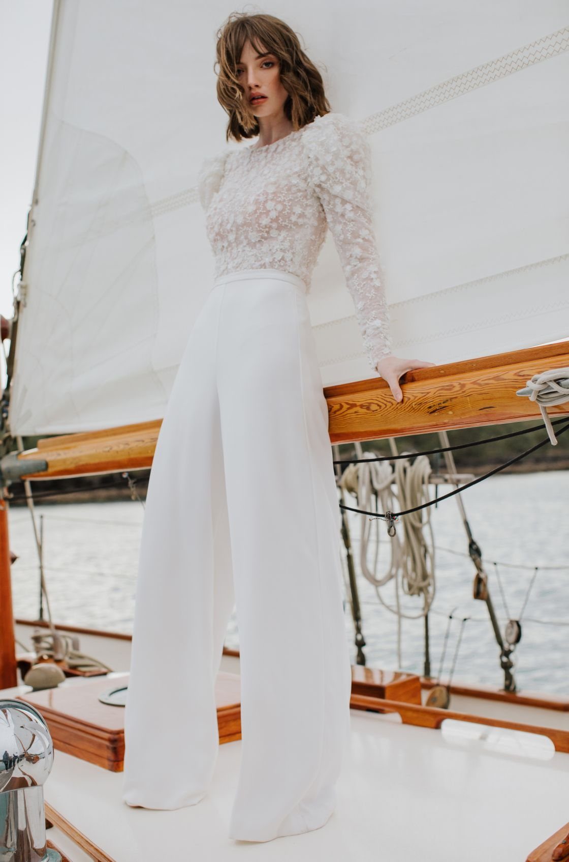 8 Civil Wedding Outfit Ideas for Brides Who Want Something Different 7 Woman in a white lace top and wide-leg trousers standing on a sailboat.