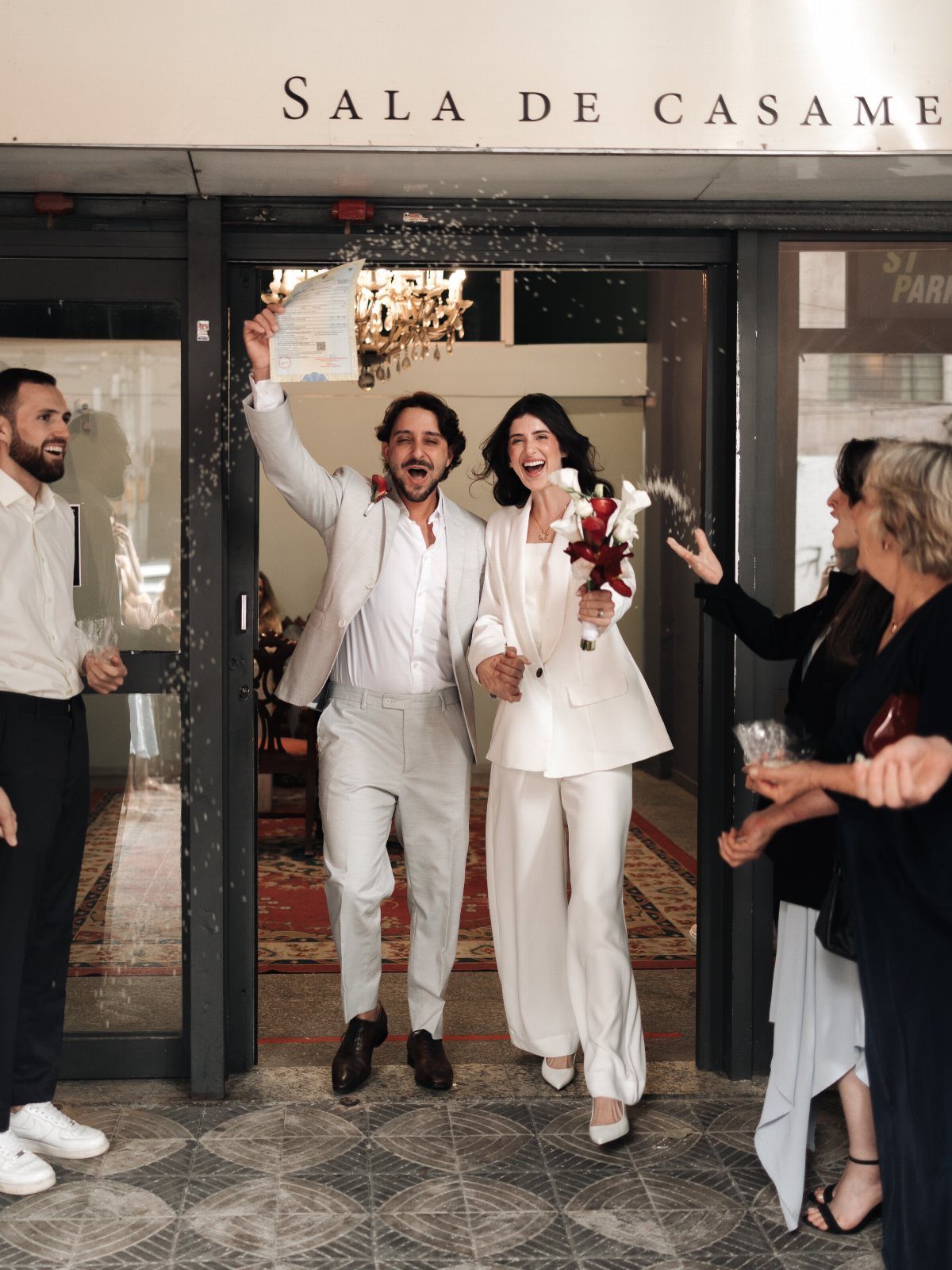 8 Civil Wedding Outfit Ideas for Brides Who Want Something Different 6 A bride in a white pantsuit and a groom in a grey suit celebrate their wedding.